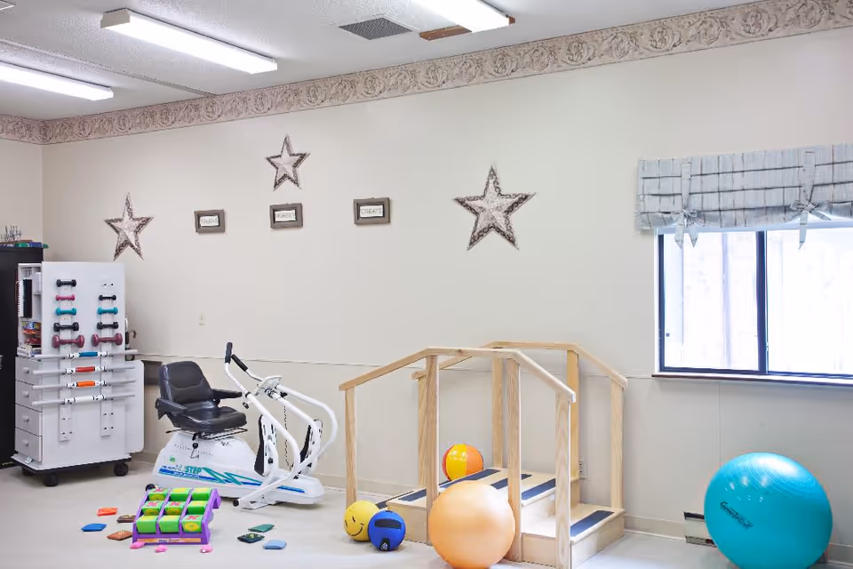A therapy or exercise room with various equipment including a stationary exercise bike, small wooden stairs with handrails, colorful therapy balls, and a rack holding small dumbbells. The room has a window with a light blue valance, beige walls decorated with star-shaped wall hangings, and fluorescent ceiling lights.