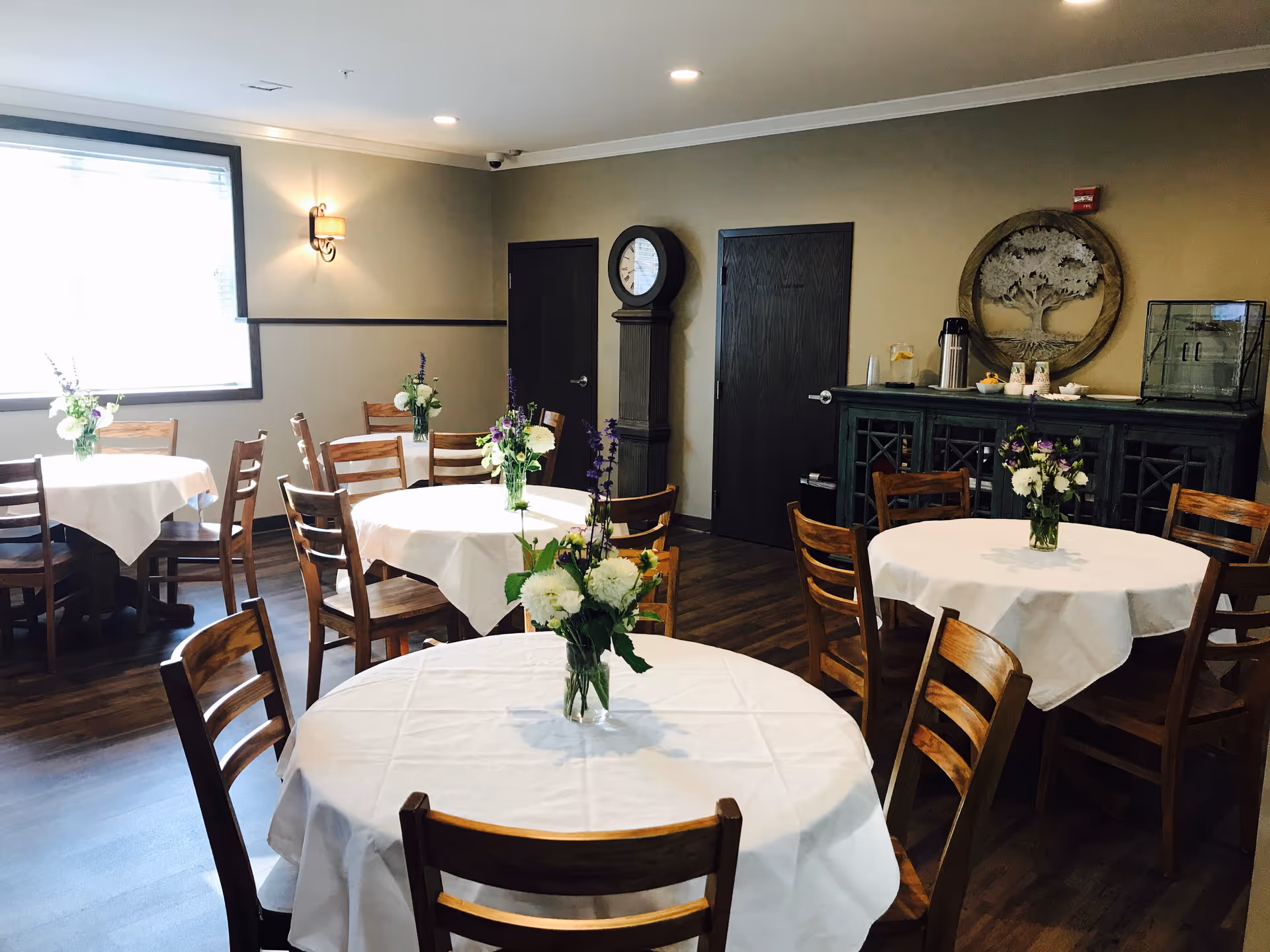 A dining room with round tables covered in white tablecloths, wooden chairs, and floral centerpieces.