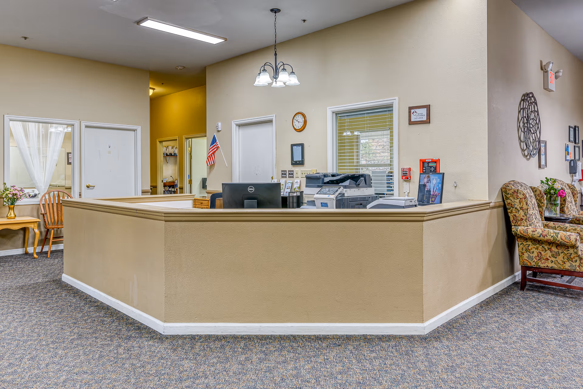 Reception desk area in an assisted living facility with a beige counter, computer monitors, printers, and an American flag. The surrounding area has beige walls, carpeted floor, floral upholstered chairs, a small wooden table with flowers, and wall decorations including a clock and framed certificates.