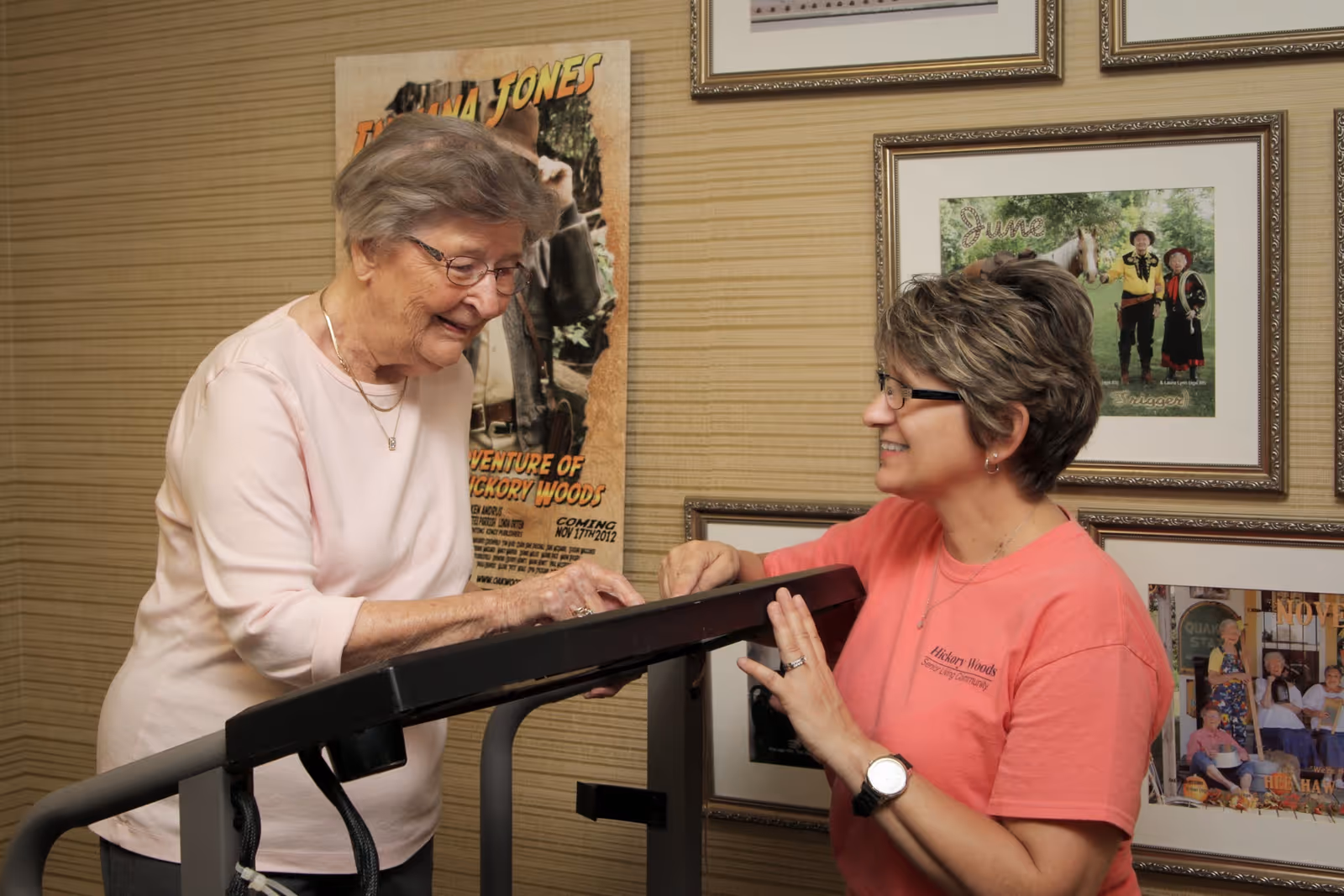 An elderly woman using a treadmill while being assisted by a staff member wearing a Hickory Woods Senior Living Community shirt in a room decorated with framed pictures and posters.