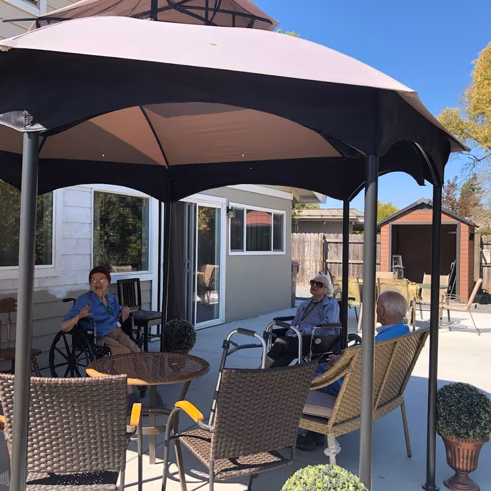 Three elderly individuals sitting outdoors under a large canopy tent in a patio area with chairs and a round table. Two of them are in wheelchairs and one is seated on a wicker chair. The background shows a building with sliding glass doors and a small shed, with a clear blue sky above.