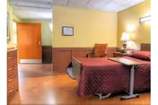 A nursing home bedroom with a hospital-style bed covered in a maroon quilt, a bedside table with a lamp and flowers, a chair, and a wooden door leading to another room. The walls are painted yellow with wooden paneling on the lower half.