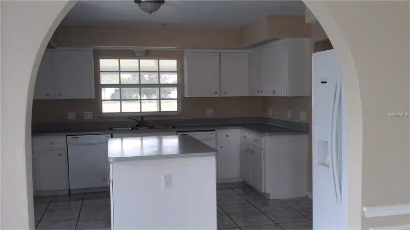 White kitchen with a center island, white cabinets, refrigerator, dishwasher and a window seen through an arched doorway.