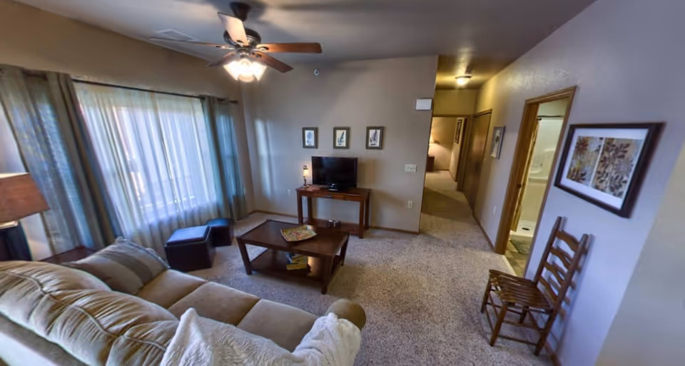 A cozy living room in a retirement community apartment featuring a beige sofa with pillows, a wooden coffee table with a decorative plate, a small TV on a wooden stand, two ottomans near a large window with sheer curtains, a ceiling fan with lights, and a hallway leading to other rooms. There is a wooden chair against the wall with framed artwork above it.