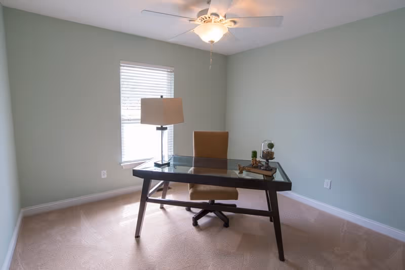 A bright, minimally furnished home office with a glass-top desk, swivel chair, table lamp, window, and ceiling fan.