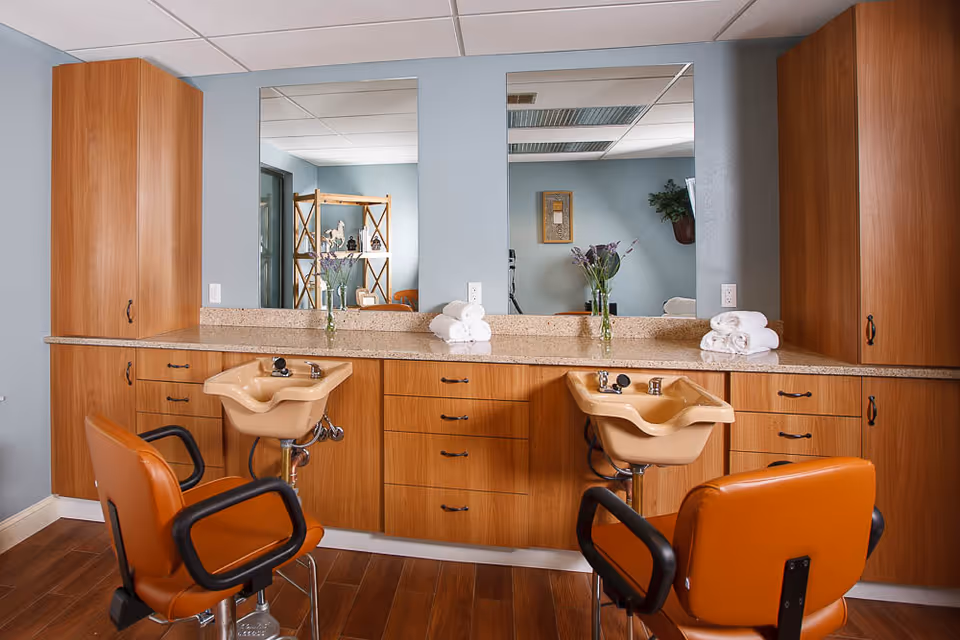 Interior view of a salon area in a senior living facility featuring two orange salon chairs in front of beige hair washing sinks. The sinks are mounted on a wooden cabinet with multiple drawers and two large mirrors above the countertop. There are rolled white towels and a vase with flowers on the countertop. The walls are painted light blue and the floor is wooden.