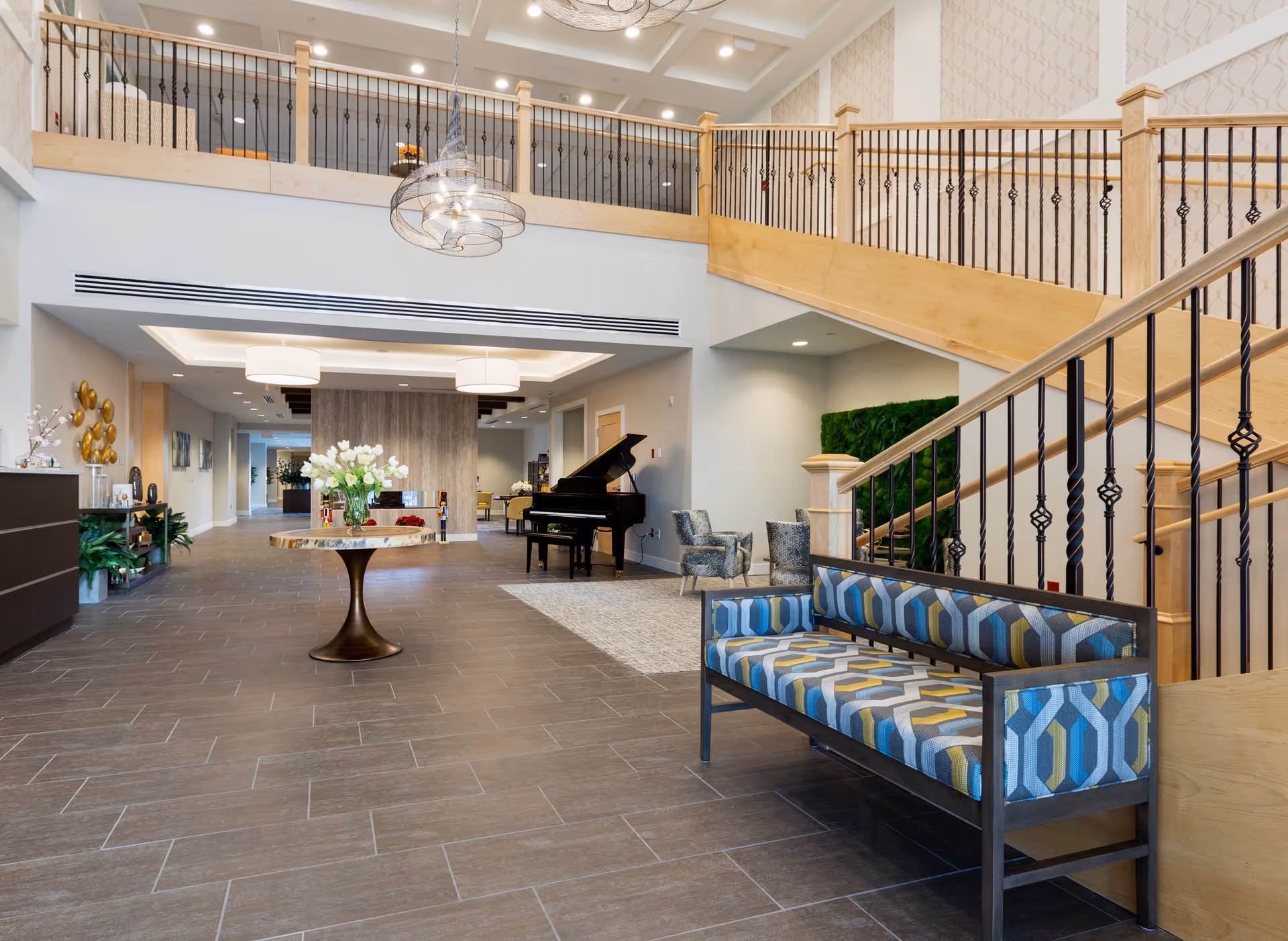 Spacious modern lobby with a central round table of flowers, patterned bench, grand piano, and a wooden staircase leading to an upper balcony.