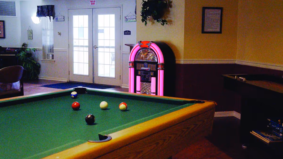 A pool table with billiard balls in a common room that includes a pink jukebox and double glass doors.