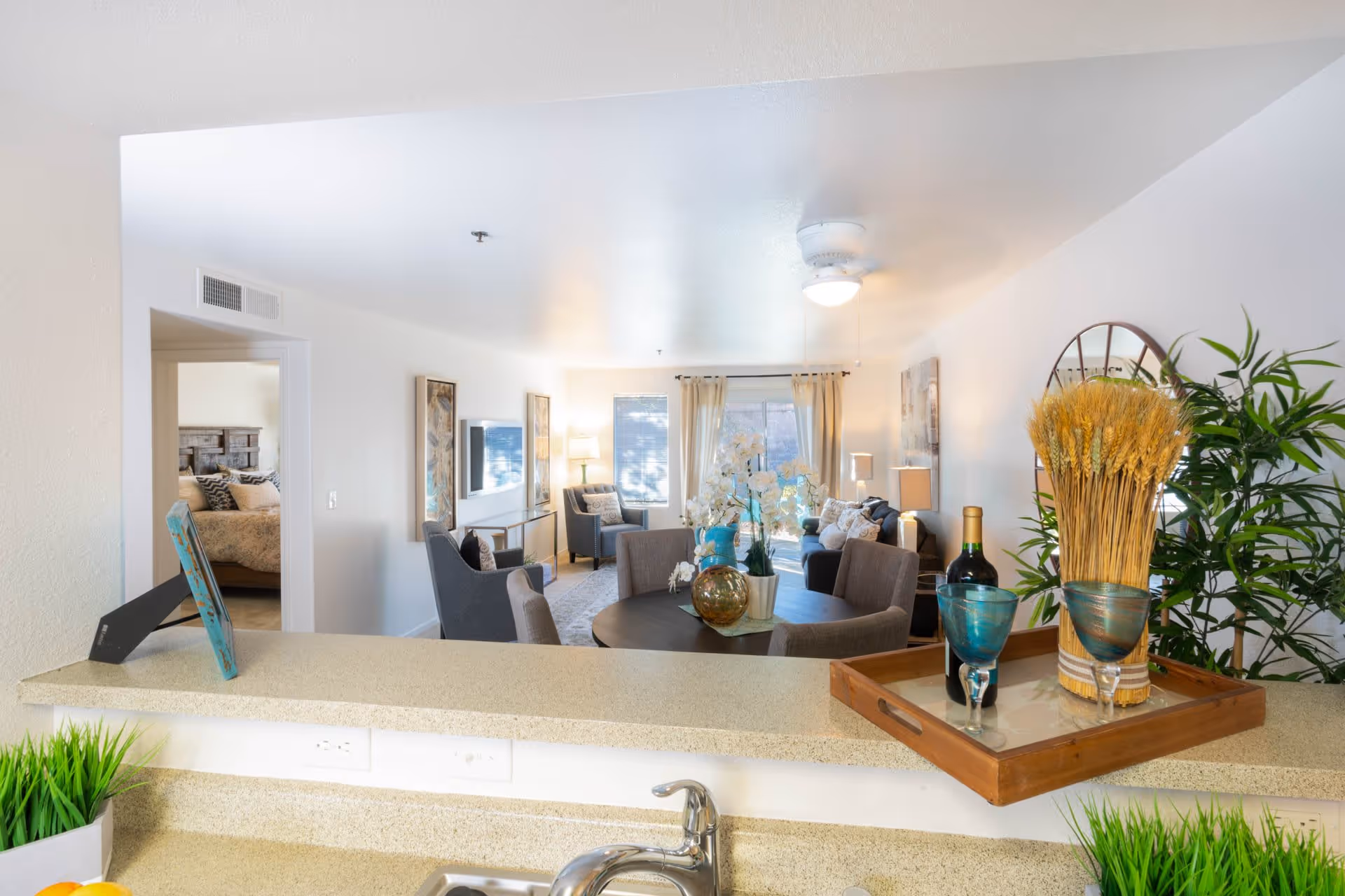 View from a kitchen counter into a bright living room with a round table, chairs, a sofa, armchairs, and large windows with curtains. Decorative items including a tray with a wine bottle, two blue glasses, and a wheat arrangement are on the counter. A bedroom with a bed is visible through an open doorway on the left.