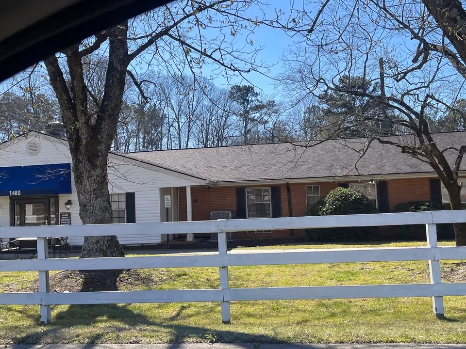 Single-story brick and white healthcare building with a blue awning, white picket fence, and trees in front.