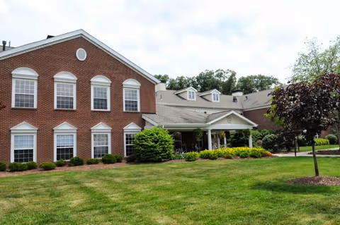 Exterior view of a senior living facility named Concordia of Cranberry, featuring a large brick building with multiple windows, a covered entrance with white columns, well-maintained green lawn, shrubs, and a tree in the foreground under a partly cloudy sky.