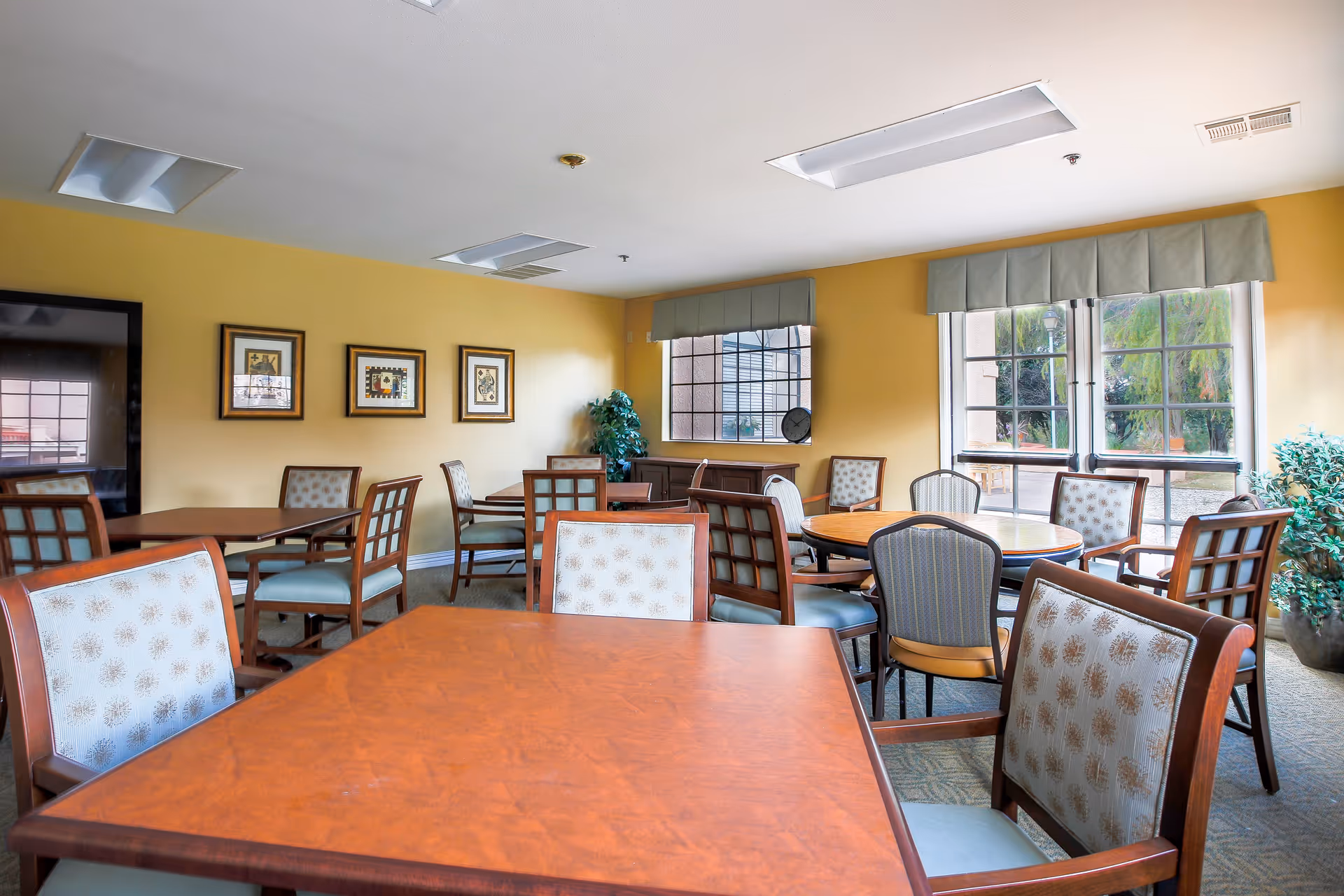 A bright dining room with multiple wooden tables and cushioned chairs arranged neatly. The walls are painted yellow and decorated with framed artwork. Large windows with green valances allow natural light to fill the room, and there are potted plants near the windows.