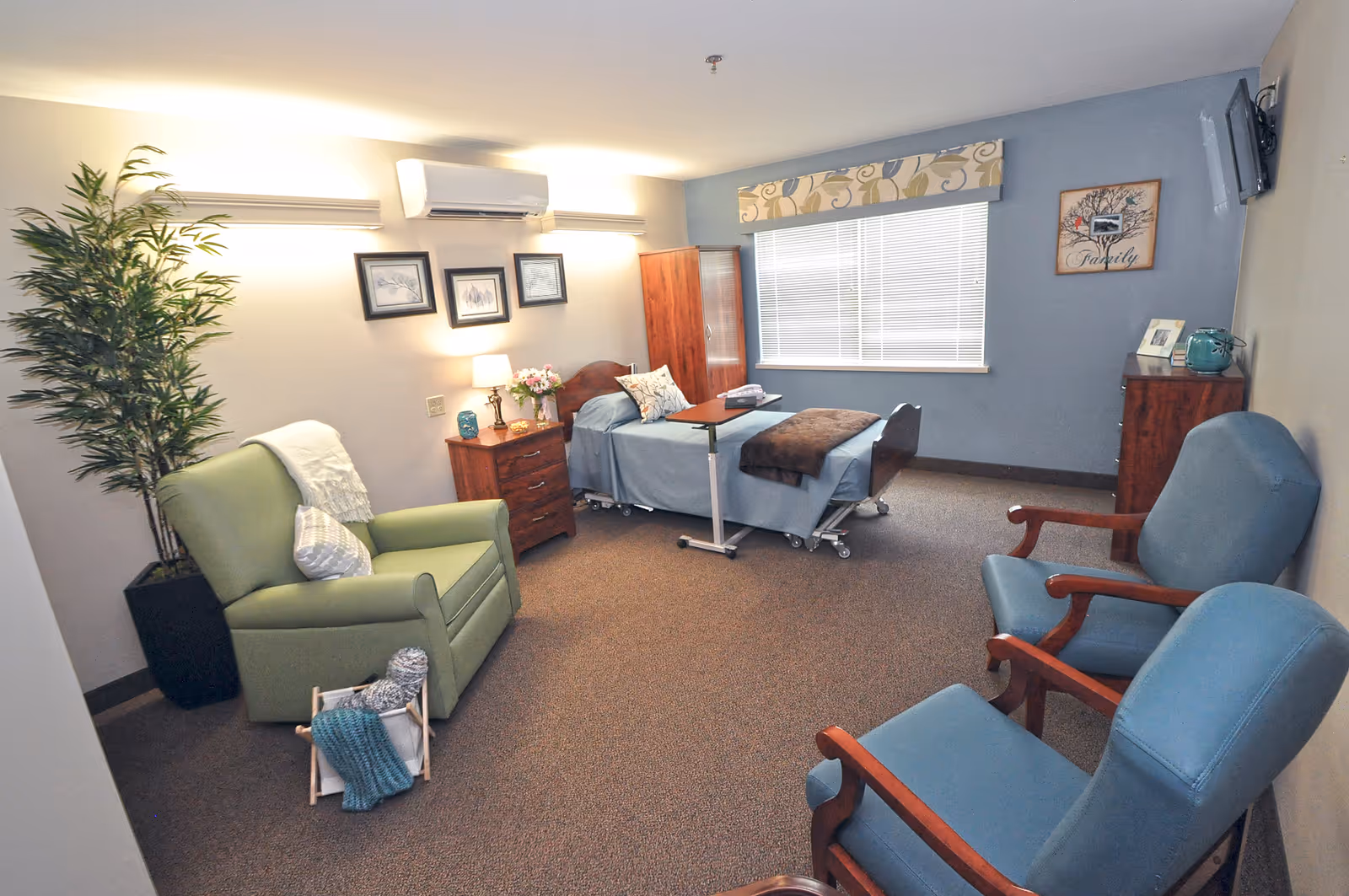 A furnished senior living facility patient room with a hospital-style bed, blue seating, and a window.
