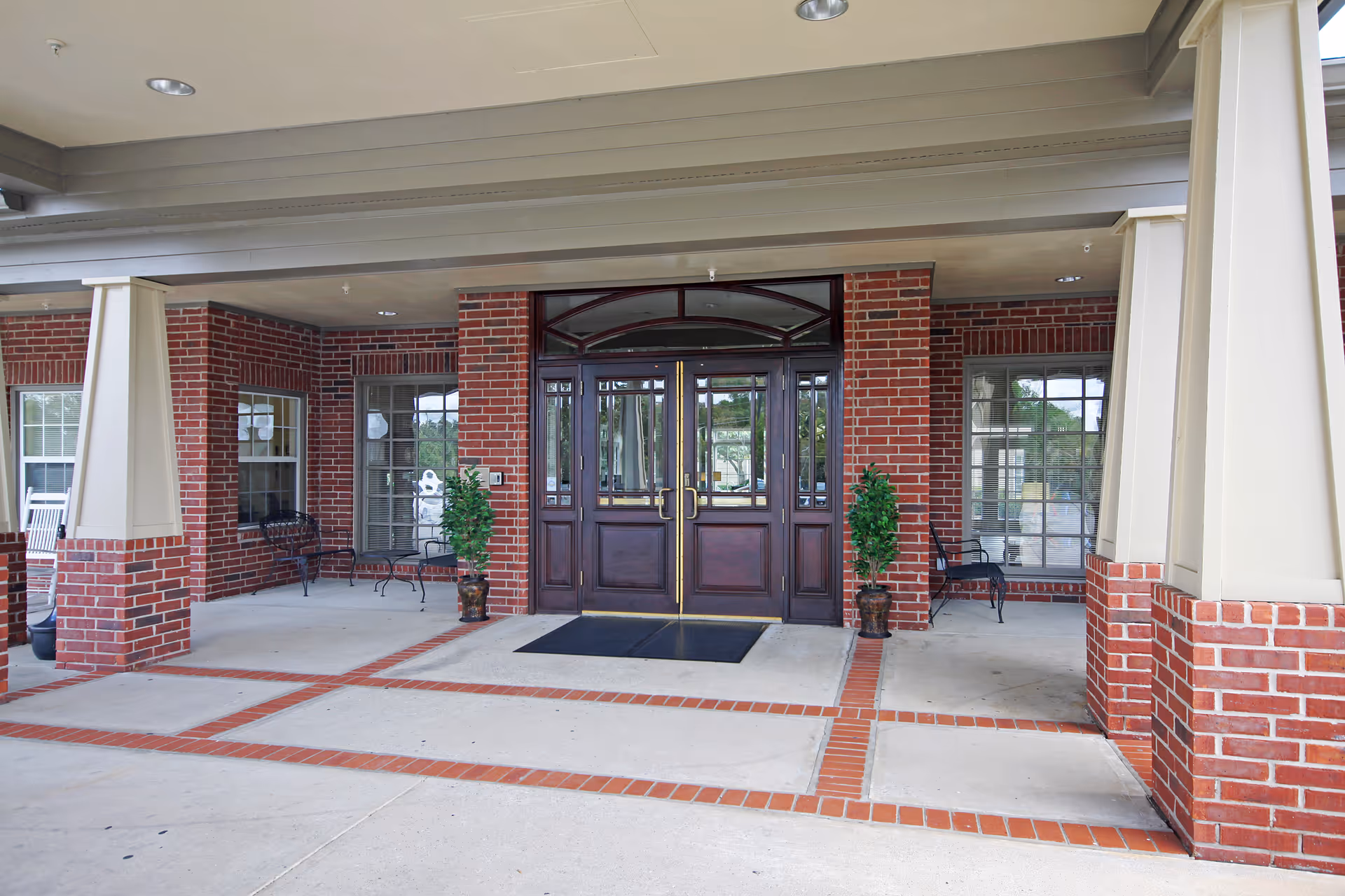 Covered brick entrance with double wooden doors, potted plants, and seating under a portico.