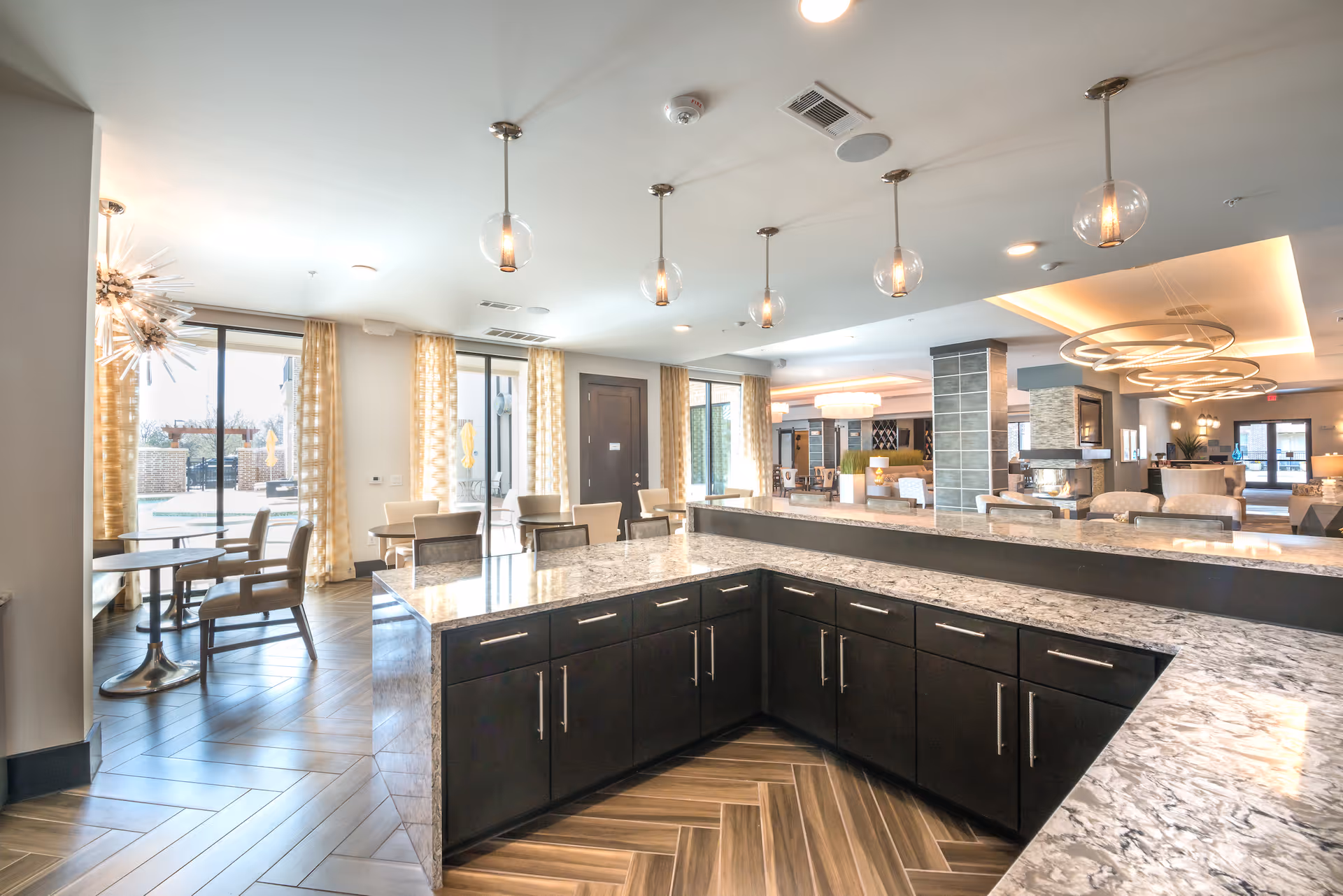 Bright open common area with a marble-topped serving island, pendant lights, and dining tables overlooking a patio.