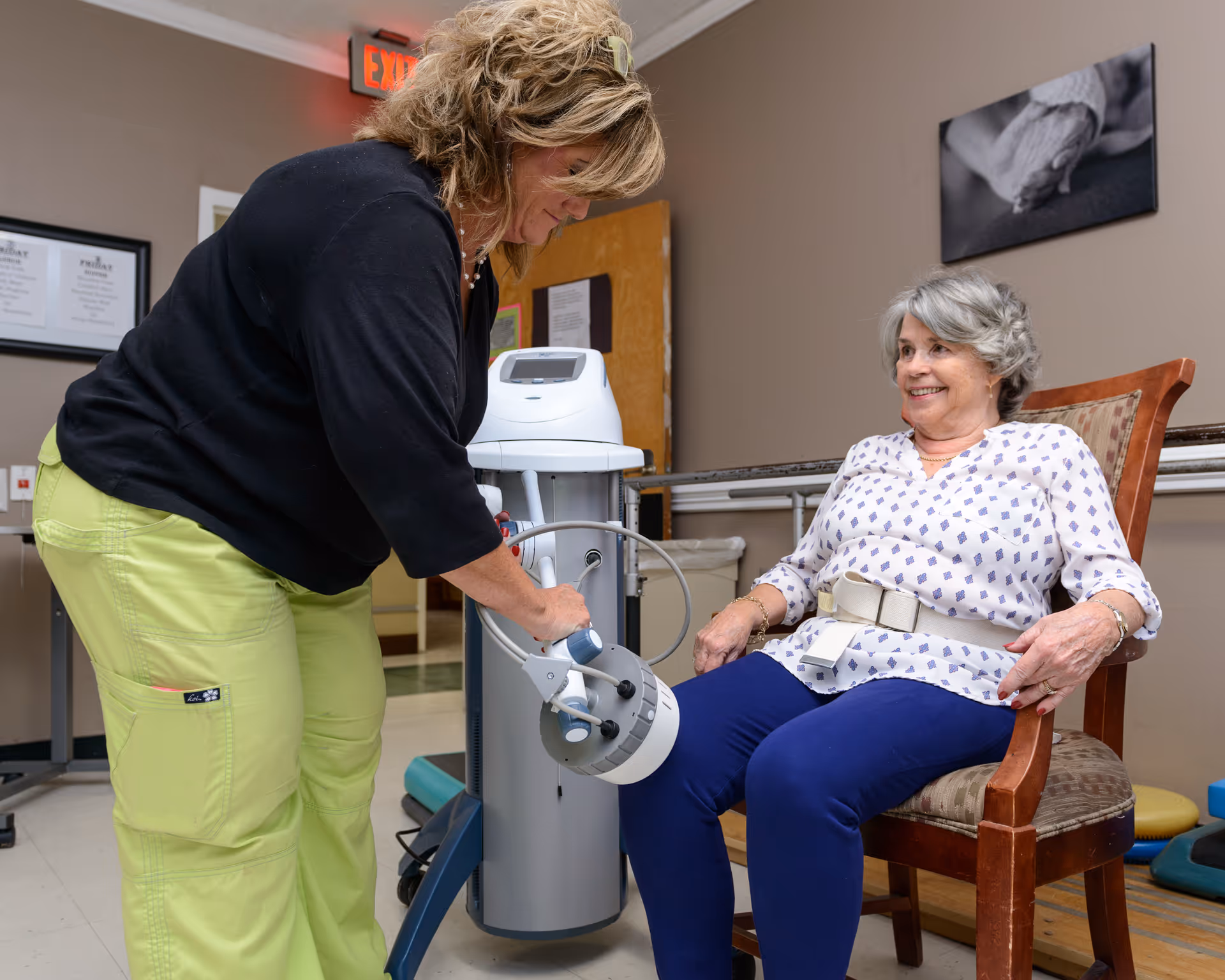 A healthcare worker is using a medical device on the knee of an elderly woman seated in a chair. The woman is smiling and wearing a white blouse with blue patterns and blue pants. The room appears to be a clinical or rehabilitation setting with medical equipment and framed documents on the wall.