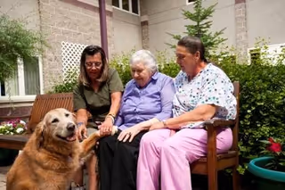 Three women sitting on a wooden bench outside near a building. One elderly woman in the center is holding hands with the two women beside her. A golden retriever dog is standing in front of them, with one woman holding the dog's paw. There are plants and flowers around them.