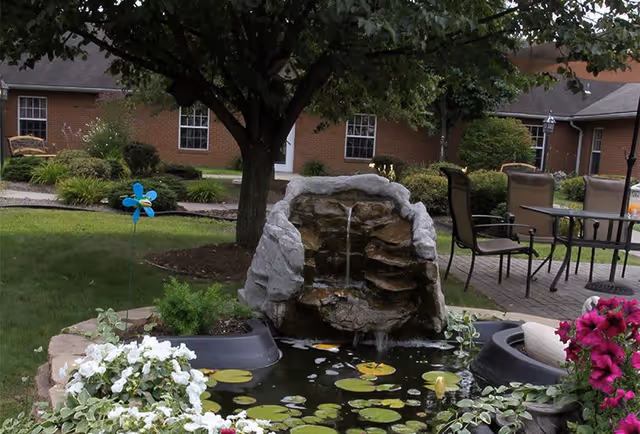 A peaceful outdoor garden area featuring a small rock waterfall flowing into a pond with lily pads, surrounded by white and purple flowers. There is a tree providing shade, a blue pinwheel decoration, and patio furniture including a table and chairs on a paved area. A brick building with windows and a door is visible in the background.