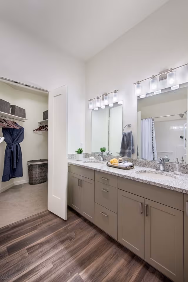 Modern bathroom with a double-sink vanity, large mirrors and lighting, and an open walk-in closet visible.