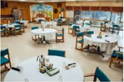 A spacious dining room with multiple round tables covered with white tablecloths, each surrounded by teal cushioned chairs. The room has large windows along one side, allowing natural light to brighten the space. The walls are decorated with a mural and some plants, creating a welcoming atmosphere.