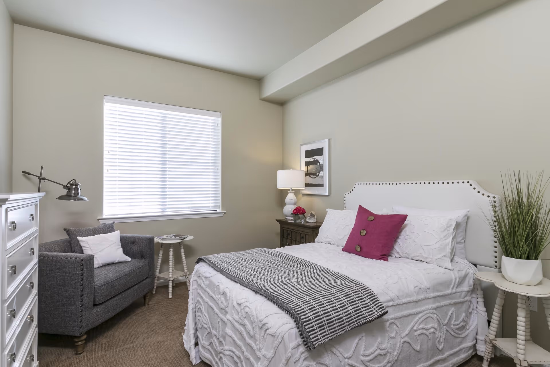 A cozy bedroom with a white bed featuring a decorative headboard, white bedding, and a magenta accent pillow. There is a gray armchair with a white pillow next to a small round white table under a window with blinds. A white dresser is on the left side, and a nightstand with a lamp, a small plant, and a framed picture is on the right side of the bed.