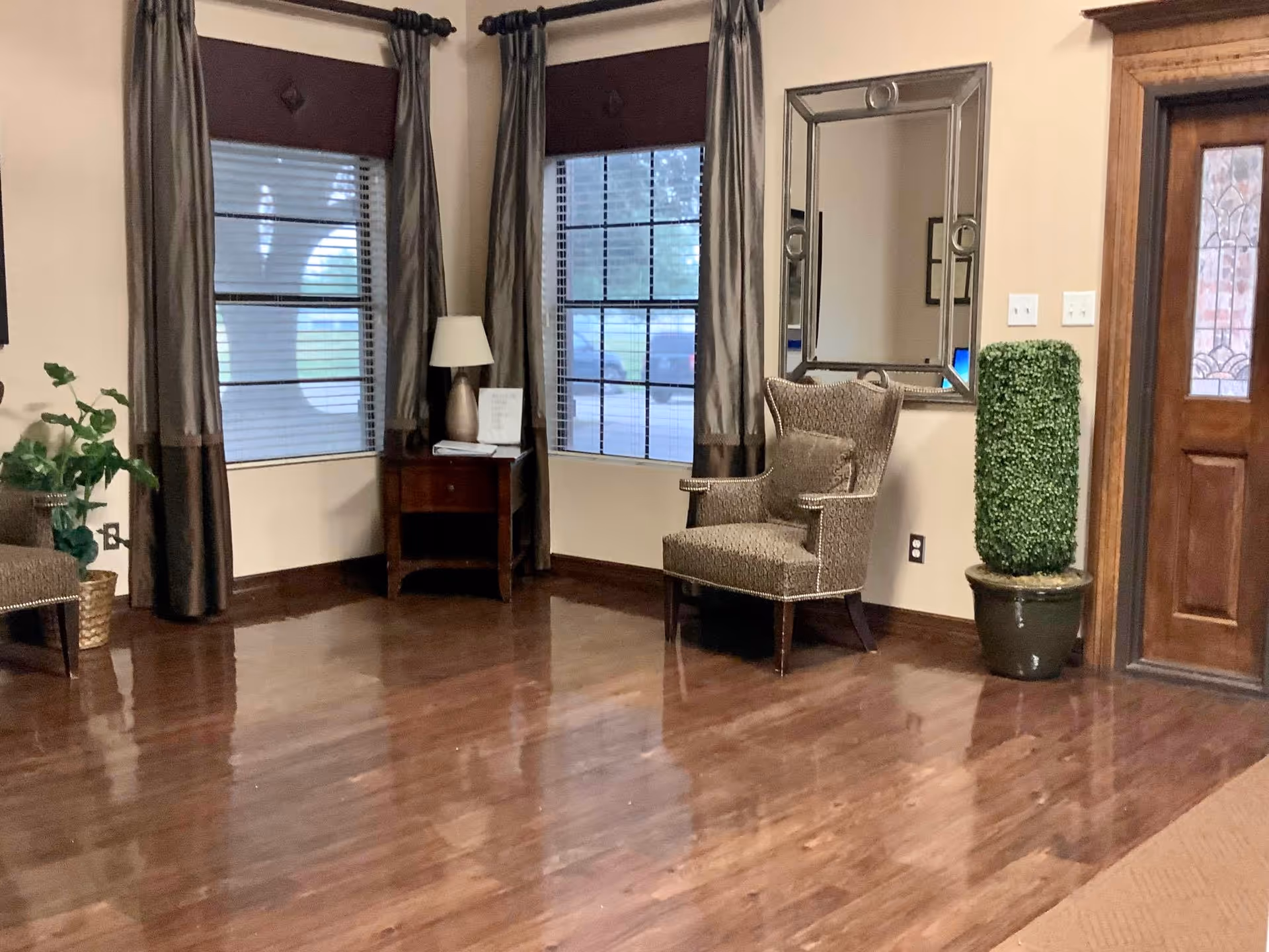A cozy interior corner of a living room with polished wooden floors, two windows with brown curtains and blinds, a wooden side table with a lamp and a small sign, a patterned armchair with a cushion, a tall potted plant, a large decorative mirror on the wall, and a wooden door with a decorative glass panel.