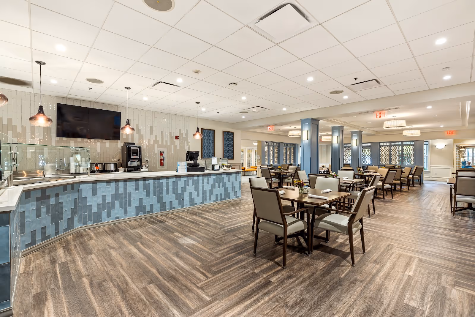 A spacious and modern dining room with multiple tables and chairs arranged neatly. The room features a long counter with a coffee machine and other appliances, pendant lights hanging from the ceiling, and a large flat-screen TV mounted on the wall. The floor has a wood-like finish, and the walls are decorated with patterned tiles and windows with decorative grilles.