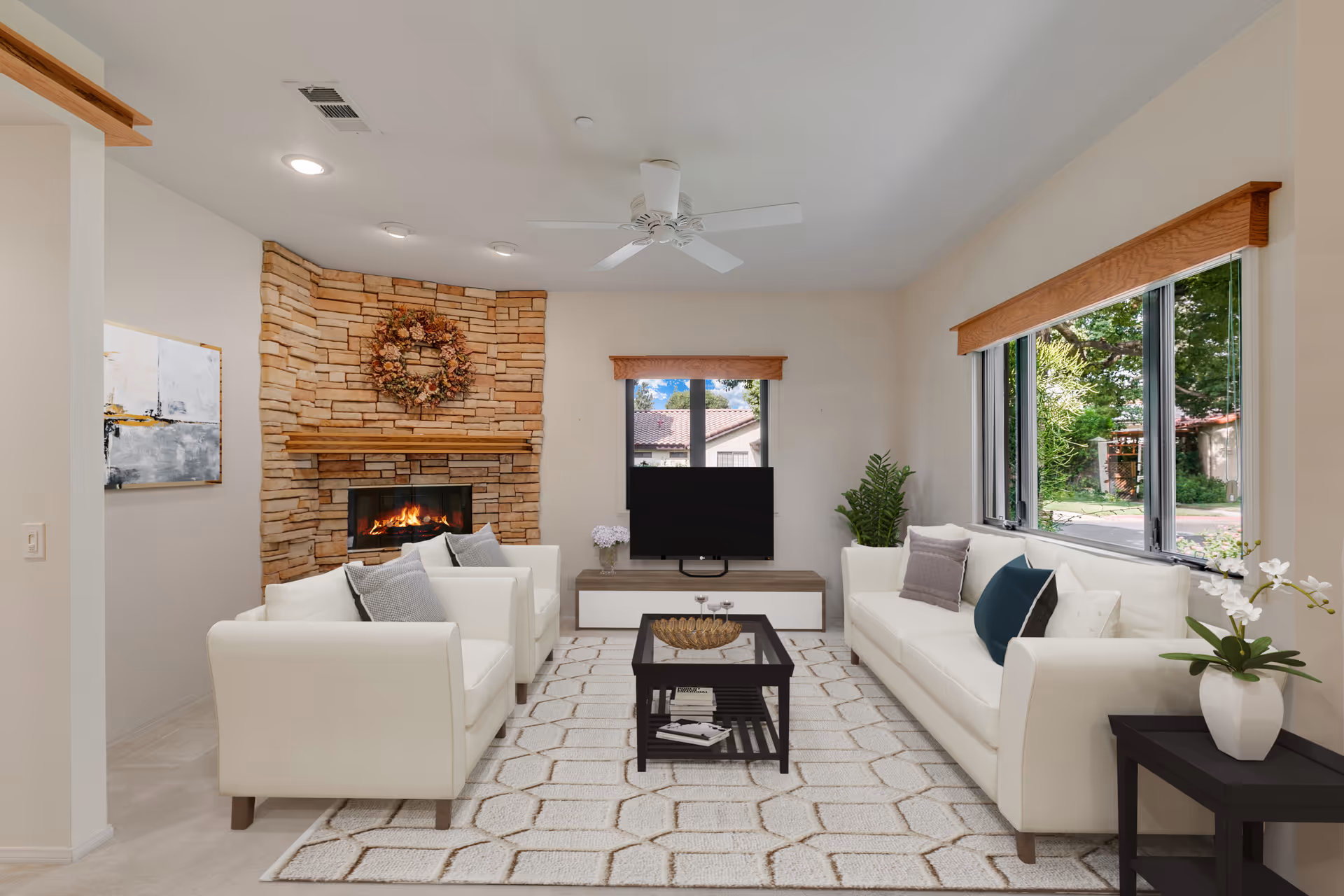 A cozy living room with a stone fireplace decorated with a floral wreath. The room has two white armchairs and a white sofa with various cushions arranged around a dark wooden coffee table on a patterned rug. A flat-screen TV sits on a low wooden stand in front of a window with wooden trim. There is a side table with a white vase and flowers next to the sofa, and large windows on the right side show greenery outside.