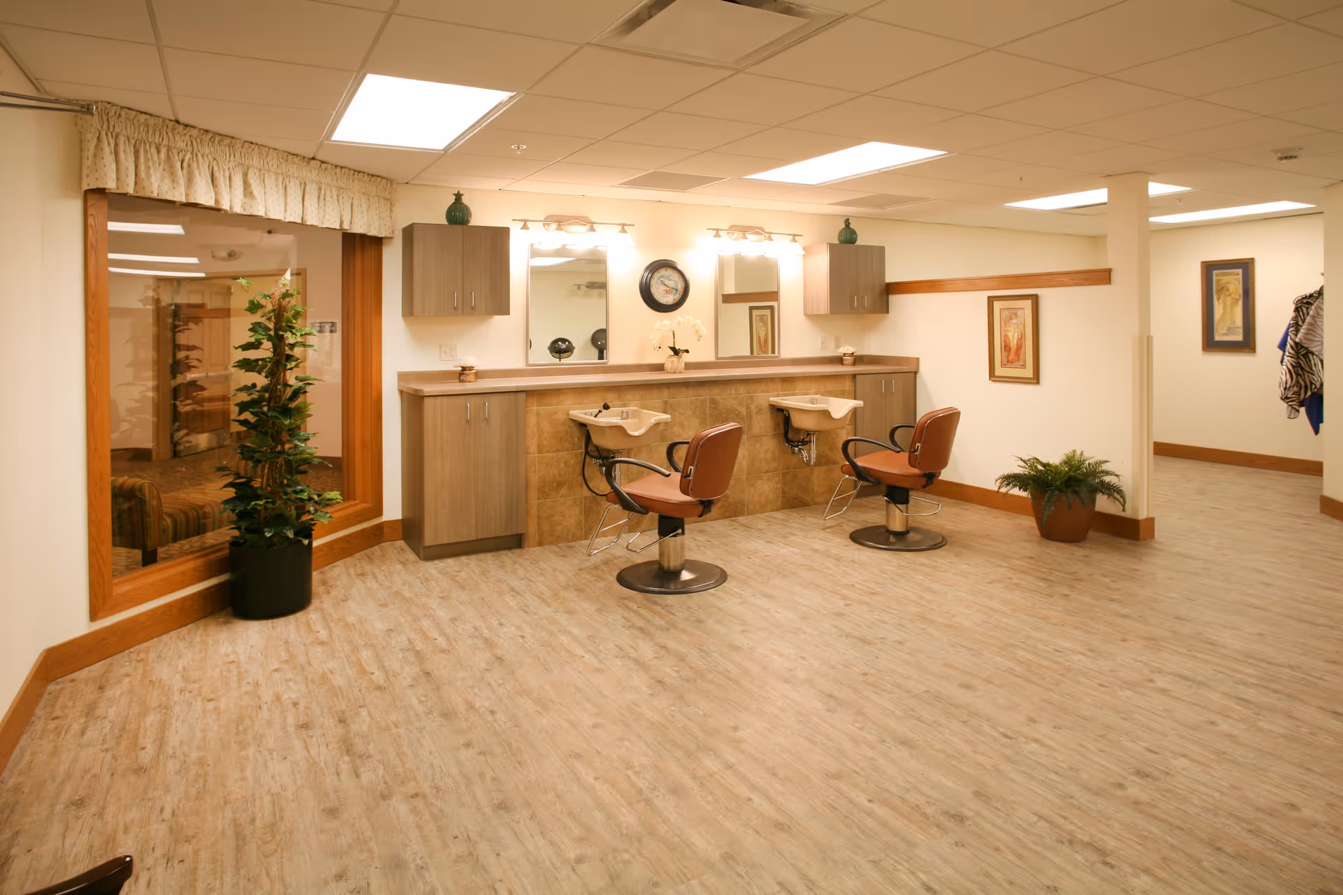 Interior view of a hair salon area in a senior living facility with two salon chairs in front of two hair washing sinks. The room has wood-look flooring, beige walls, a large window with a plant in front, and framed artwork on the walls. There are cabinets and mirrors above the sinks with lighting fixtures.