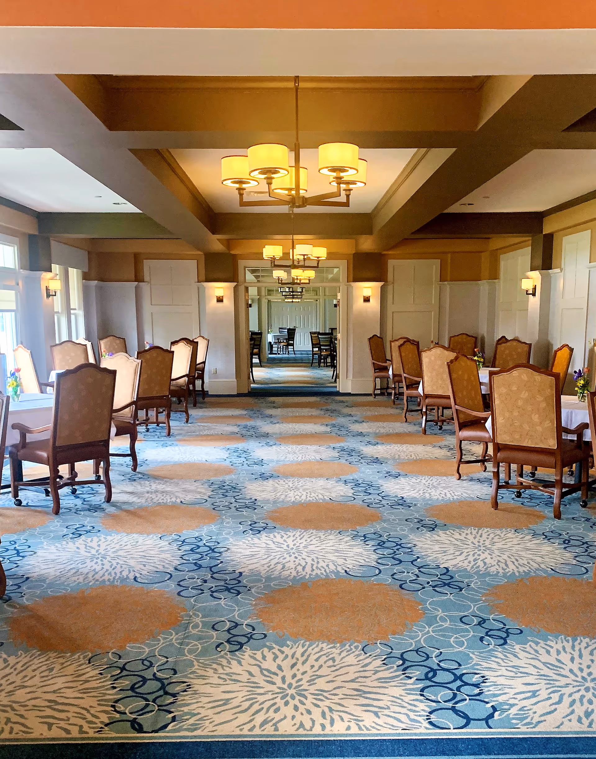 Spacious formal dining room with patterned carpet, multiple tables and upholstered chairs under chandeliers.