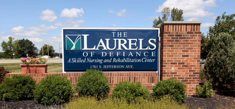 A large blue and white sign for The Laurels of Defiance, a skilled nursing and rehabilitation center, located at 1701 S. Jefferson Ave. The sign is mounted on a brick base with green bushes and a flower pot with colorful flowers nearby. The background shows a partly cloudy sky and some trees.