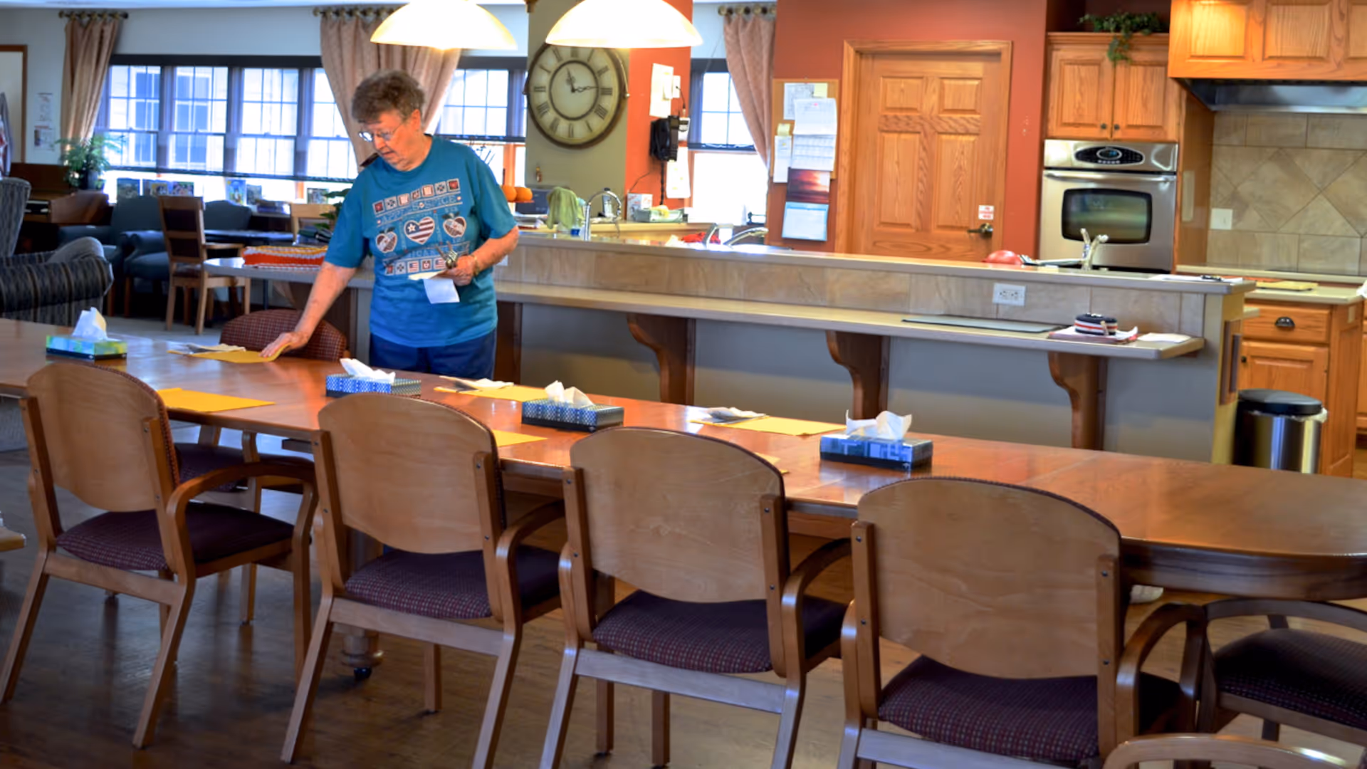 An older woman arranges papers and tissue boxes on a long communal dining table in a dining area with an open kitchen behind her.