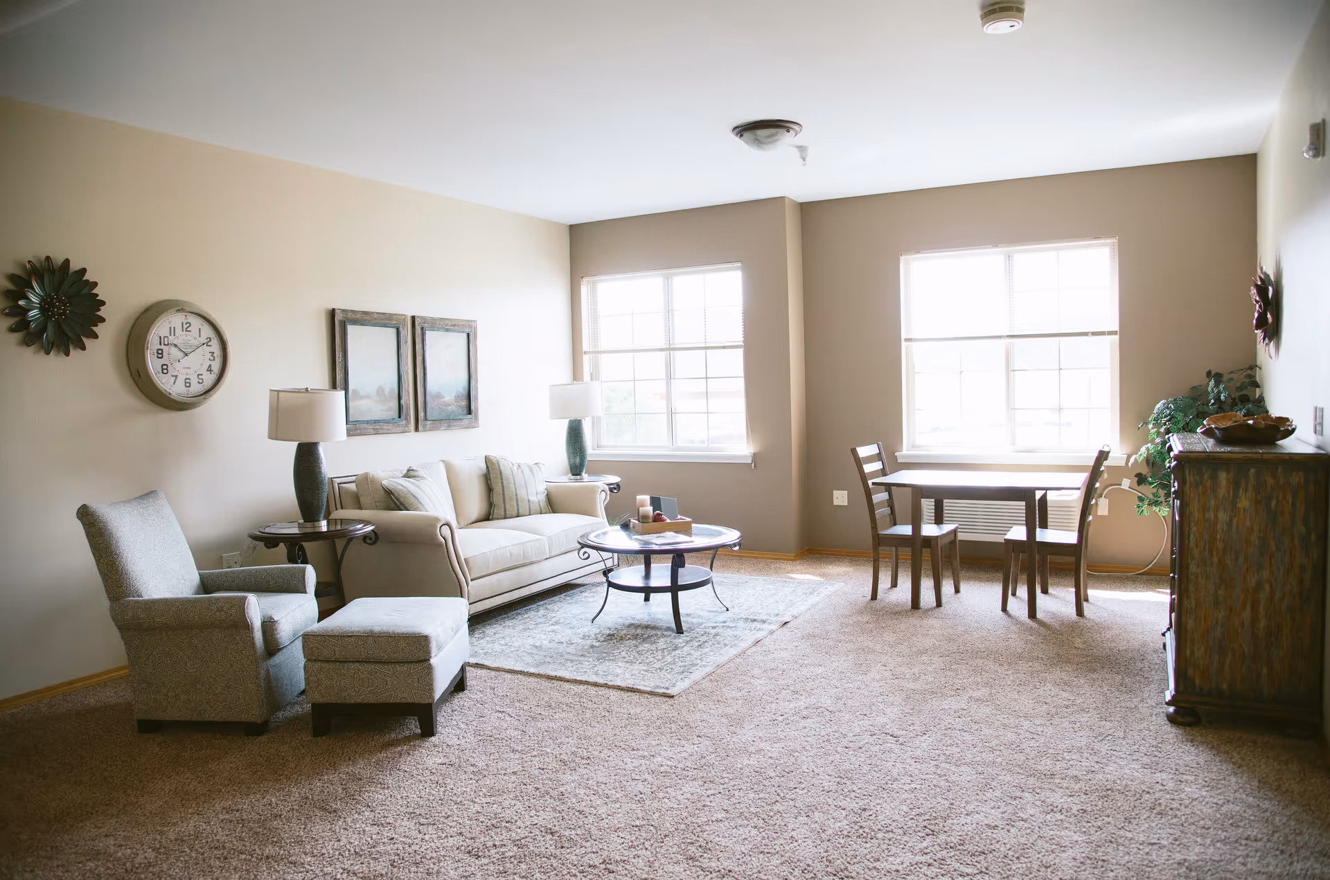 Bright furnished living room with a sofa, armchair and ottoman, coffee table, and a small dining table by two large windows.