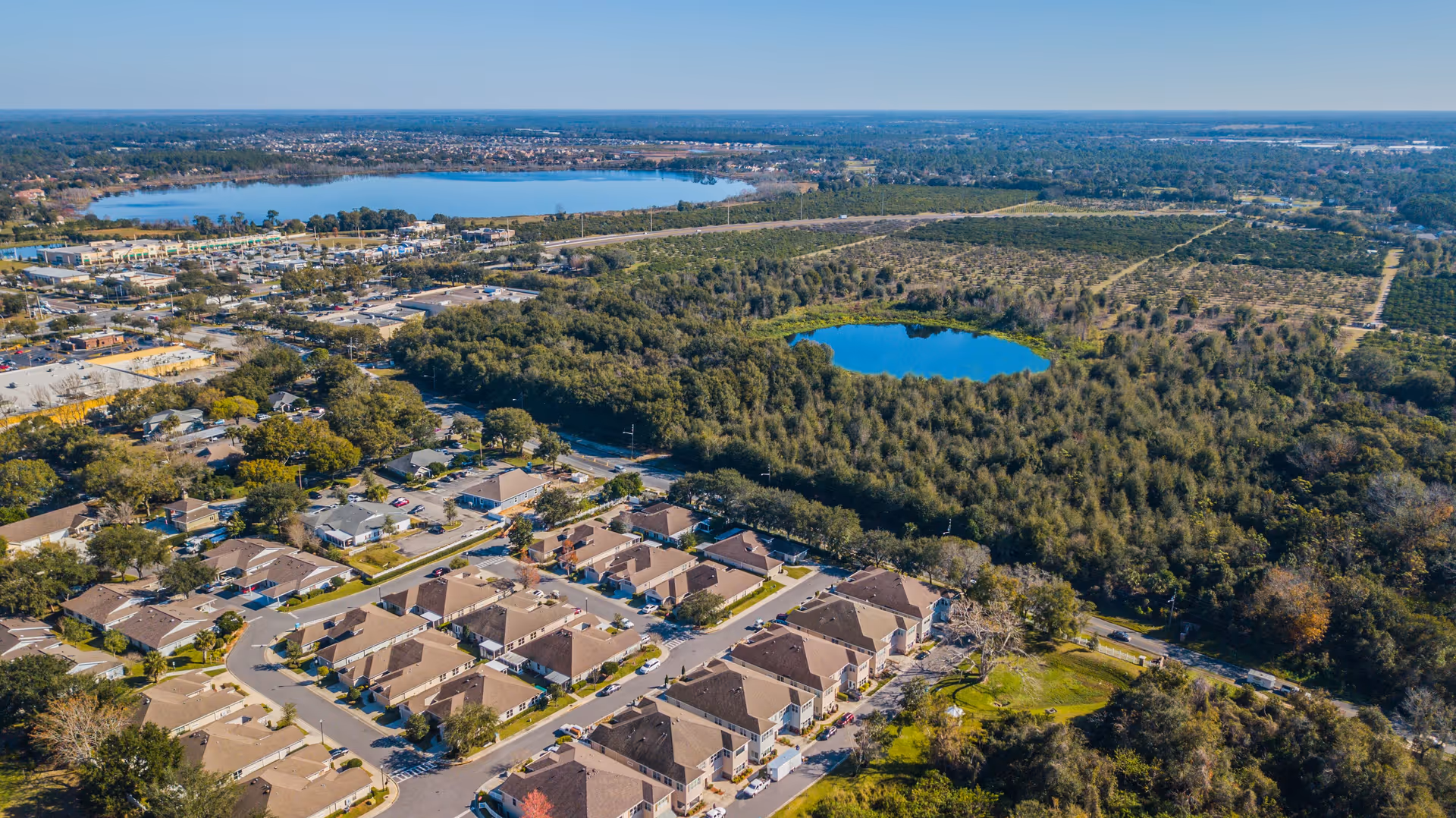 Aerial view of a residential community with rows of houses, surrounded by trees and greenery. There are two visible lakes in the background, with a mix of forested and open land extending to the horizon.
