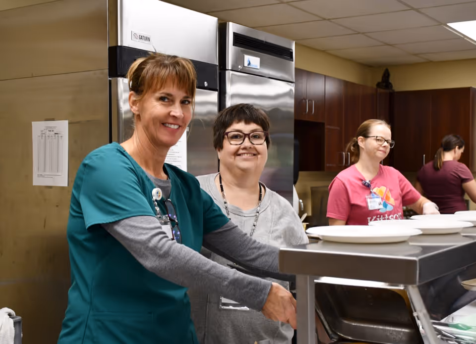 Four women working in a kitchen area. One woman in green scrubs is smiling at the camera while washing dishes. Another woman in gray stands beside her smiling. Two other women in pink shirts are working in the background near a counter with plates.