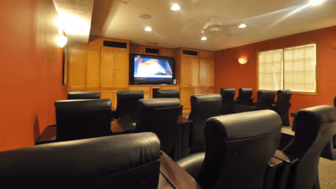 A small theater room with multiple rows of black leather recliner chairs facing a flat-screen TV mounted on a wooden cabinet wall. The room has warm lighting, red walls, and a window with blinds on the right side.
