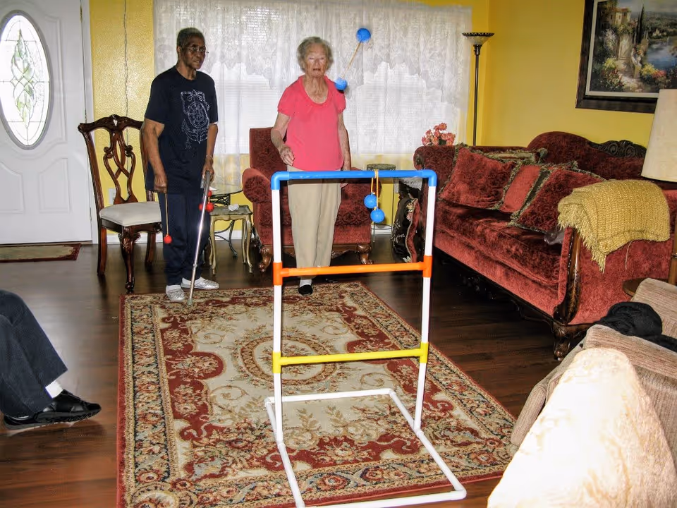 Two elderly women playing ladder toss game in a living room with wooden floor, a patterned area rug, red upholstered furniture, and yellow walls. One woman is holding a cane and the other is standing behind the game frame.