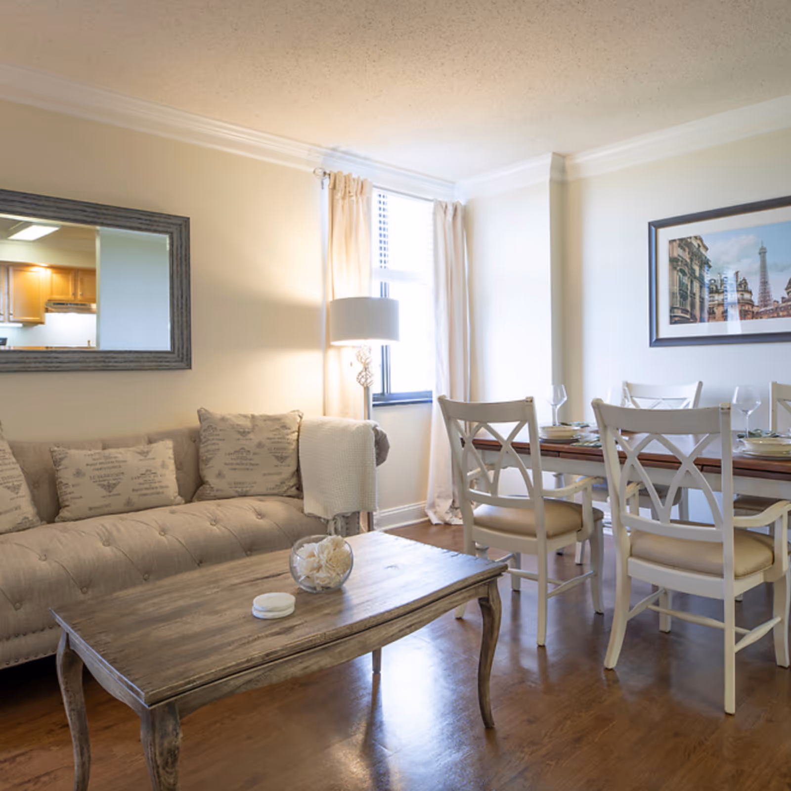 A cozy living and dining area featuring a beige tufted sofa with decorative pillows, a wooden coffee table with a glass bowl centerpiece, a dining table set with four white chairs and place settings, a floor lamp near a window with cream curtains, and a framed picture on the wall.
