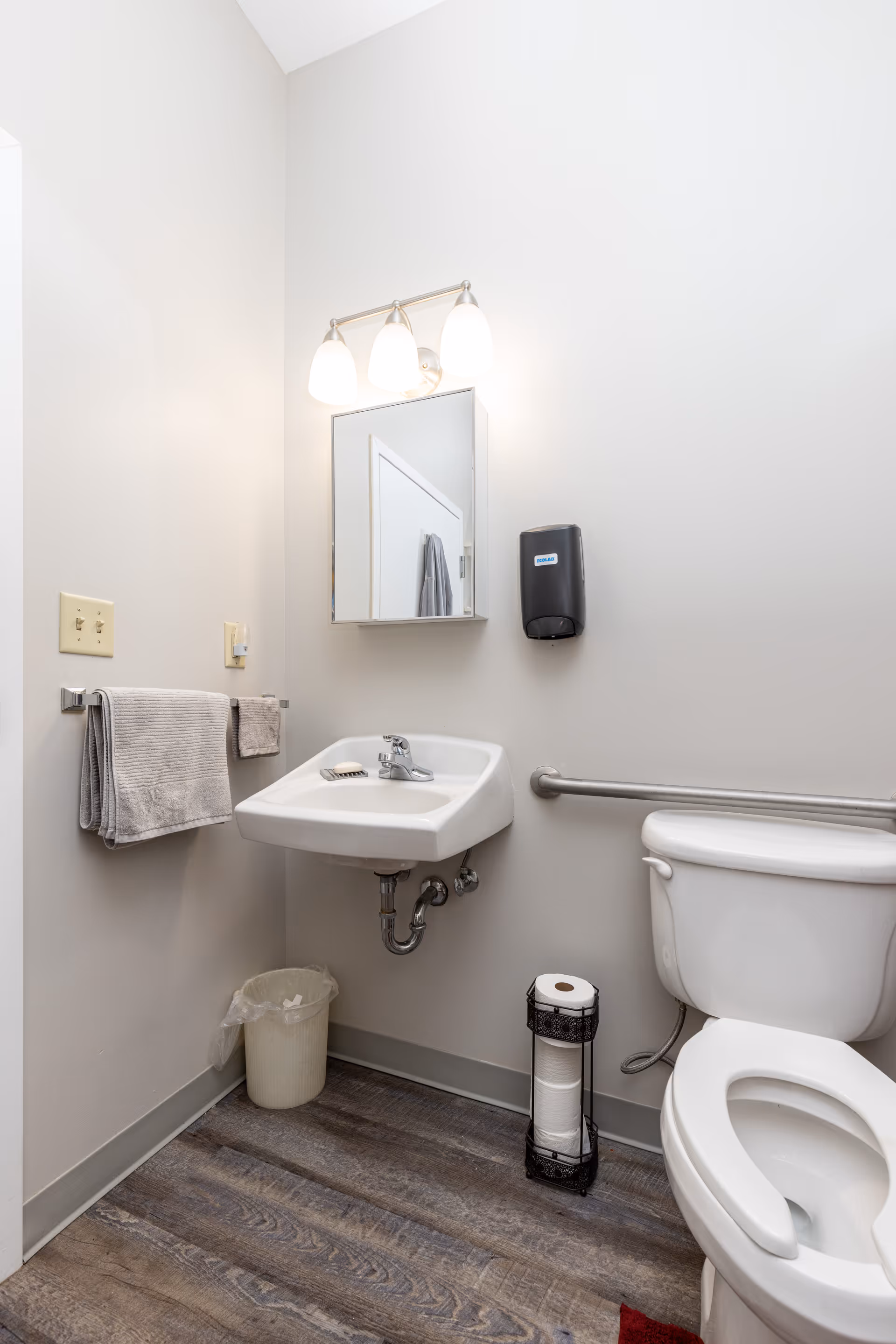 A small bathroom with a white toilet, a wall-mounted sink with a soap dish, a mirror with three lights above it, a towel rack with two towels, a trash can, and a toilet paper holder with multiple rolls. The walls are light-colored and the floor has a wood-like finish.