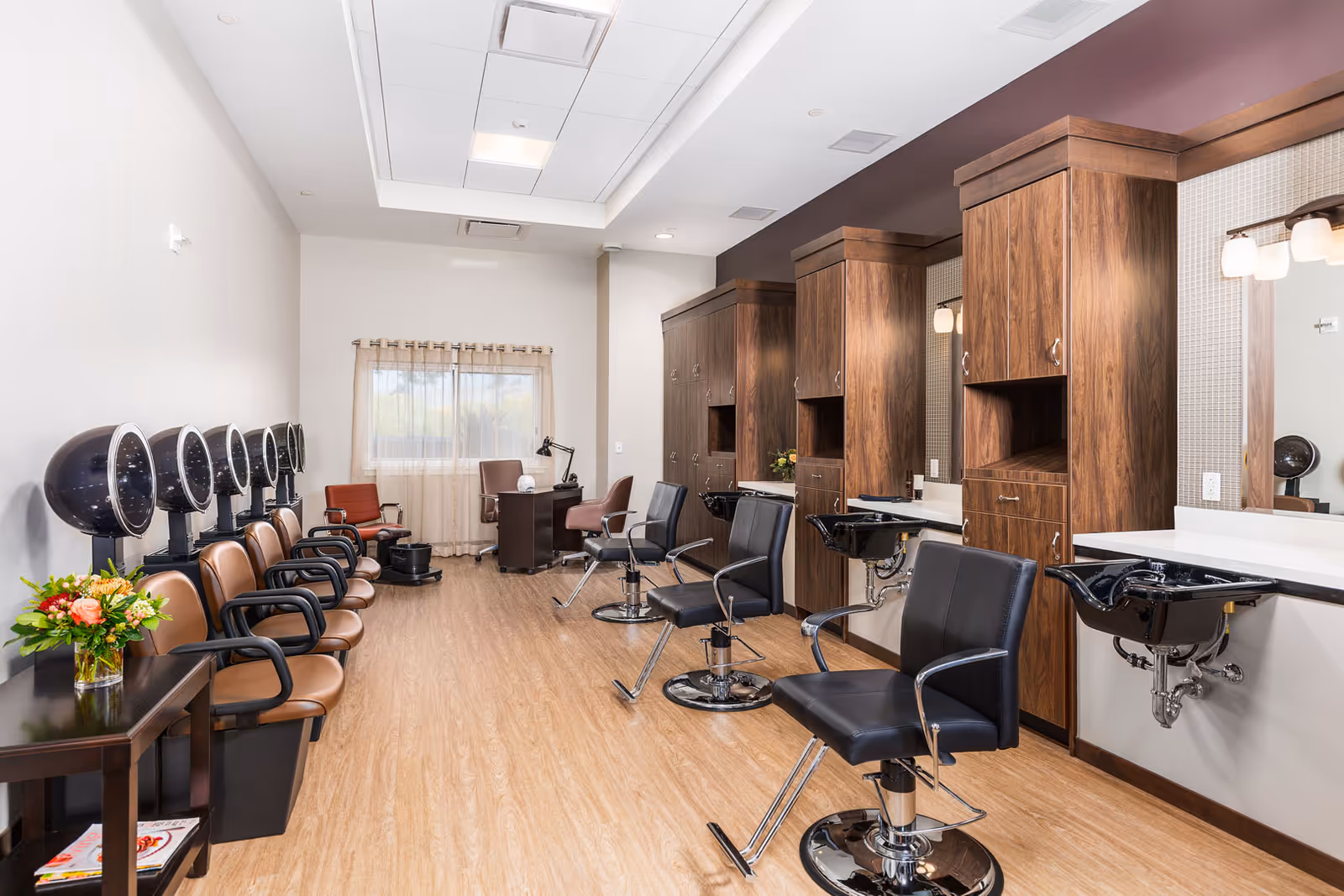 A modern salon room with several black salon chairs in front of wooden cabinets and sinks for hair washing. On the left side, there are multiple brown chairs with black hair dryers above them. A small table with a flower arrangement and magazines is also visible. The room has light wood flooring, white walls, and a window with sheer curtains at the far end.