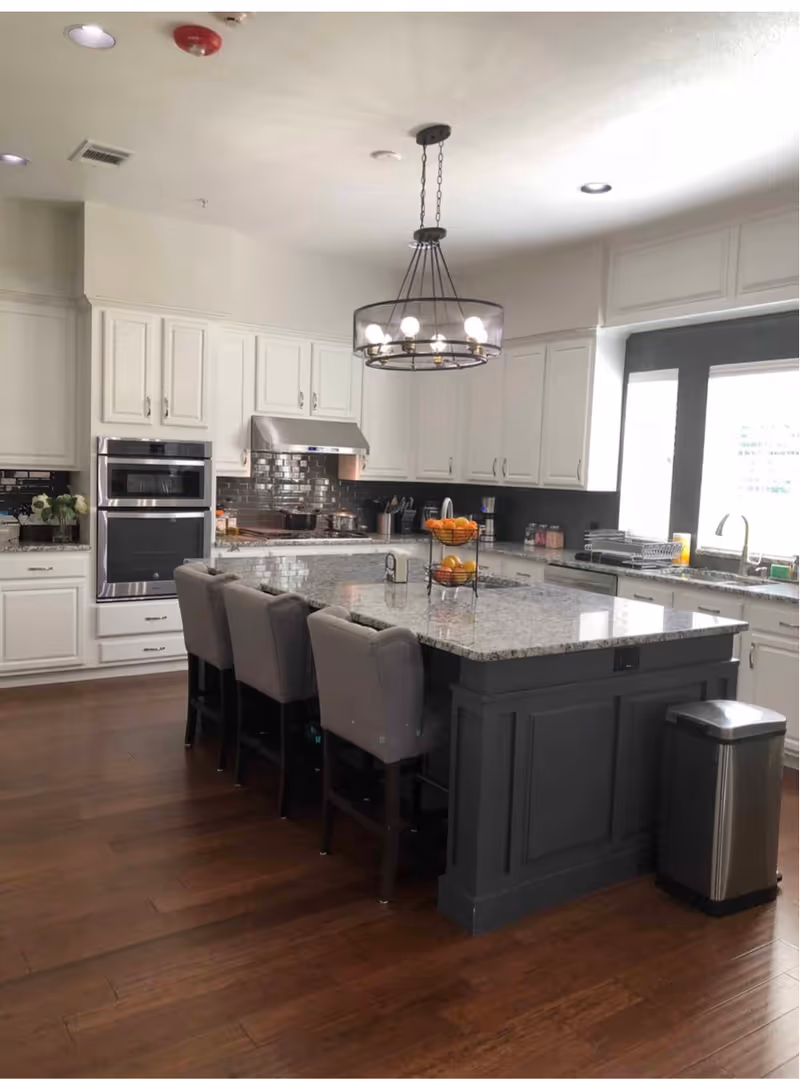 A modern kitchen with white cabinets, a large granite island with four gray upholstered chairs, stainless steel appliances including an oven and range hood, a fruit stand with oranges and apples on the island, and a window letting in natural light.