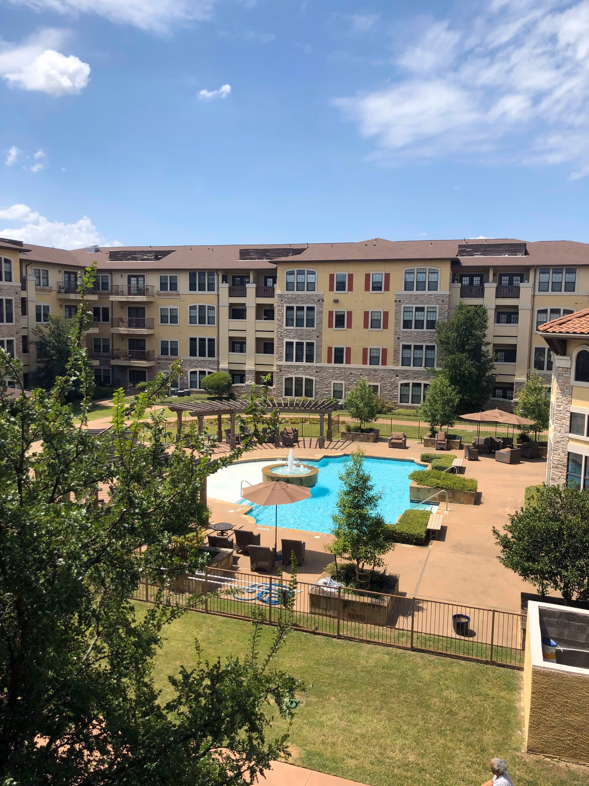 Outdoor view of a senior living facility courtyard with a swimming pool, lounge chairs, umbrellas, and a pergola. The multi-story building surrounds the courtyard, and there are trees and green grass areas under a partly cloudy blue sky.