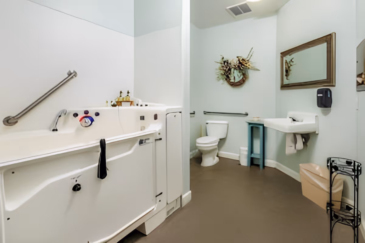 A clean, accessible bathroom with a walk-in bathtub equipped with grab bars, a toilet, a wall-mounted sink with a towel underneath, a mirror above the sink, a small blue table, a decorative wreath on the wall, and a trash bin in the corner.