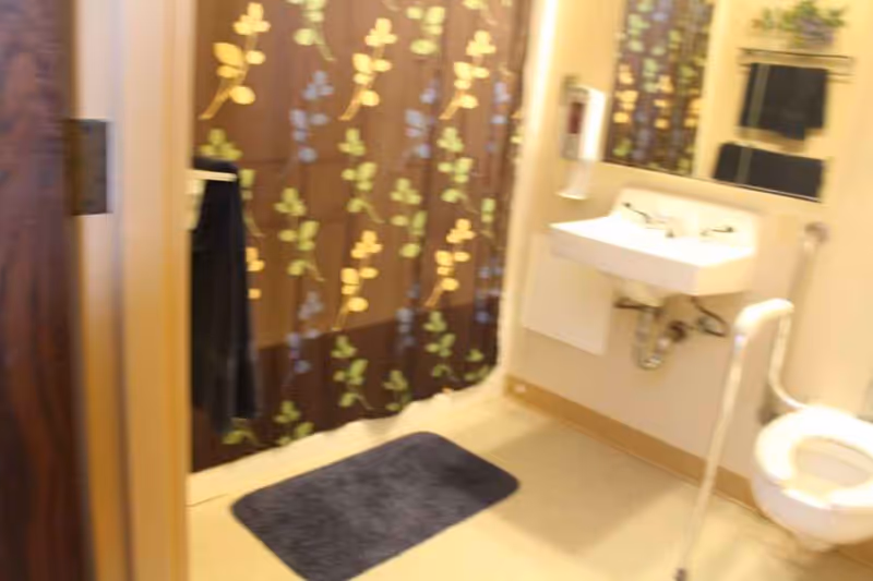 A bathroom with a floral patterned shower curtain, a wall-mounted sink, a toilet with a support rail, a dark bath mat on the floor, and a towel hanging on a rack.