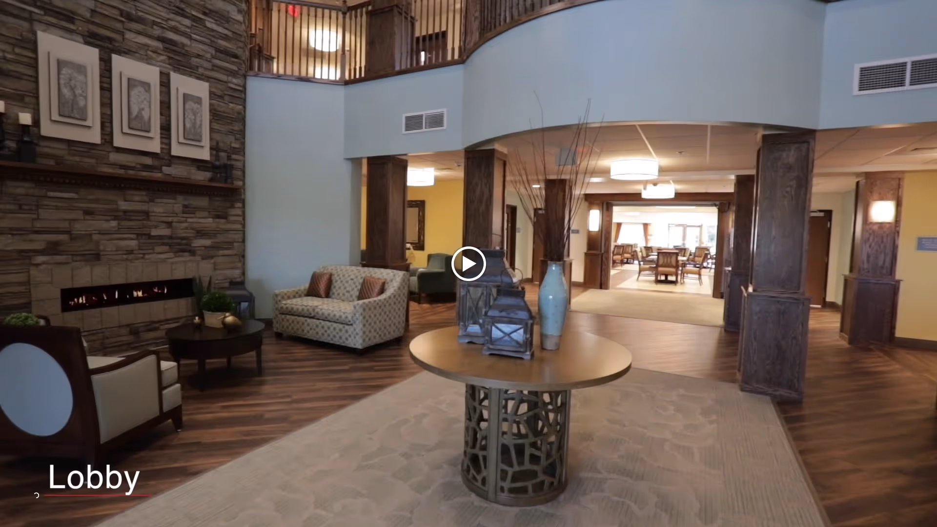 Spacious two-story lobby with seating around a fireplace, a central round table with decorative vases, and a view into a dining area.