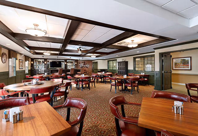 A spacious dining room with multiple wooden tables and red leather chairs arranged neatly. The room features a patterned carpet, coffered ceiling with recessed lighting, and a bar area with stools and a TV mounted on the wall. The walls are painted in green and beige tones with framed artwork and windows.