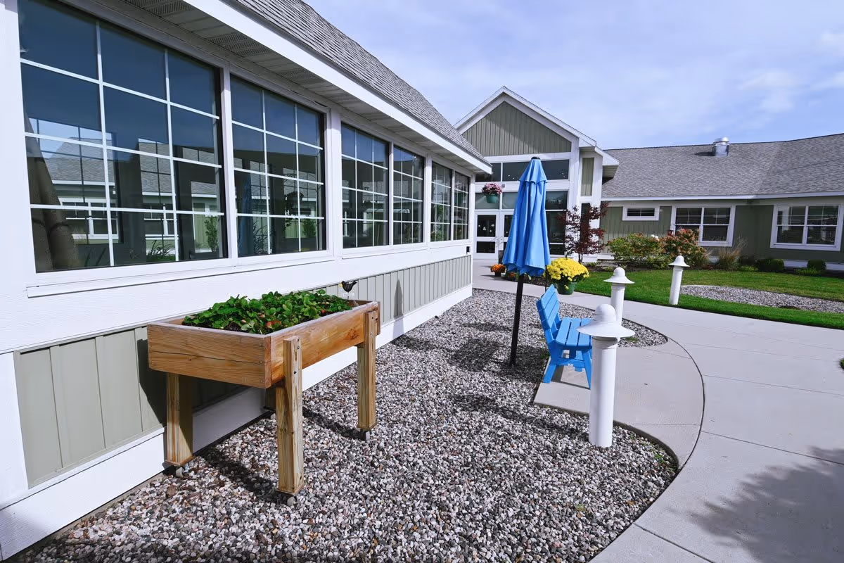 Outdoor area of The Cortland Holland Meadows facility featuring a gravel bed with a wooden planter box filled with green plants, a blue bench with a closed blue umbrella, white pathway lights, and a building with large windows and a peaked roof in the background under a partly cloudy sky.