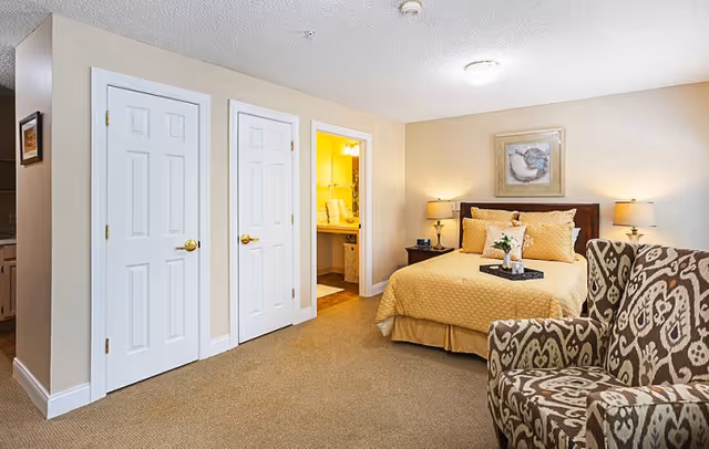 A cozy furnished bedroom with a bed, bedside tables and lamps, a patterned armchair, two white closet doors, and an open doorway to a bathroom.