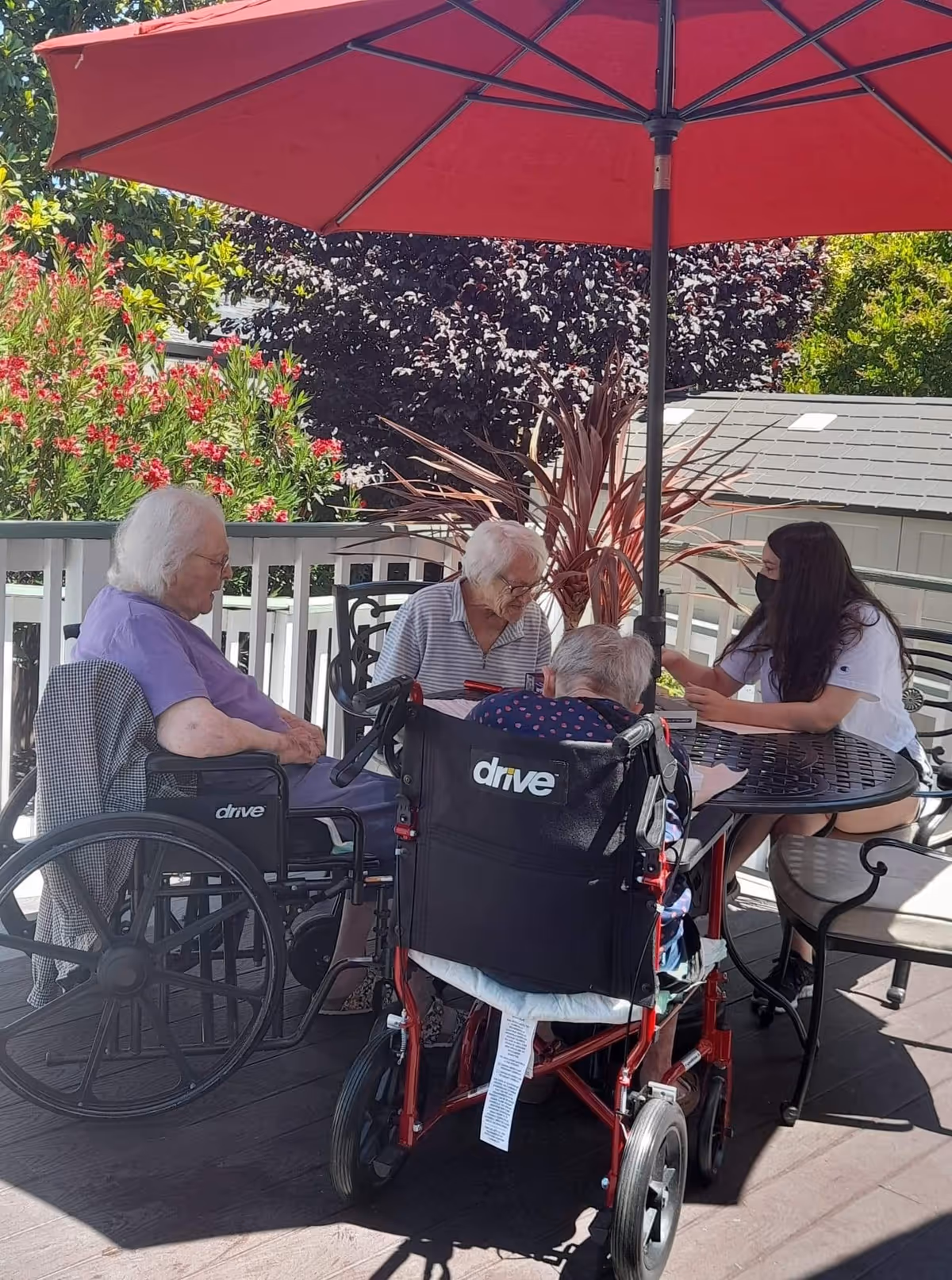 Three elderly individuals, two in wheelchairs, sitting around a round outdoor table under a large red umbrella. A younger woman wearing a mask is seated with them, engaging in conversation. The setting is a patio area with plants and a white fence in the background.