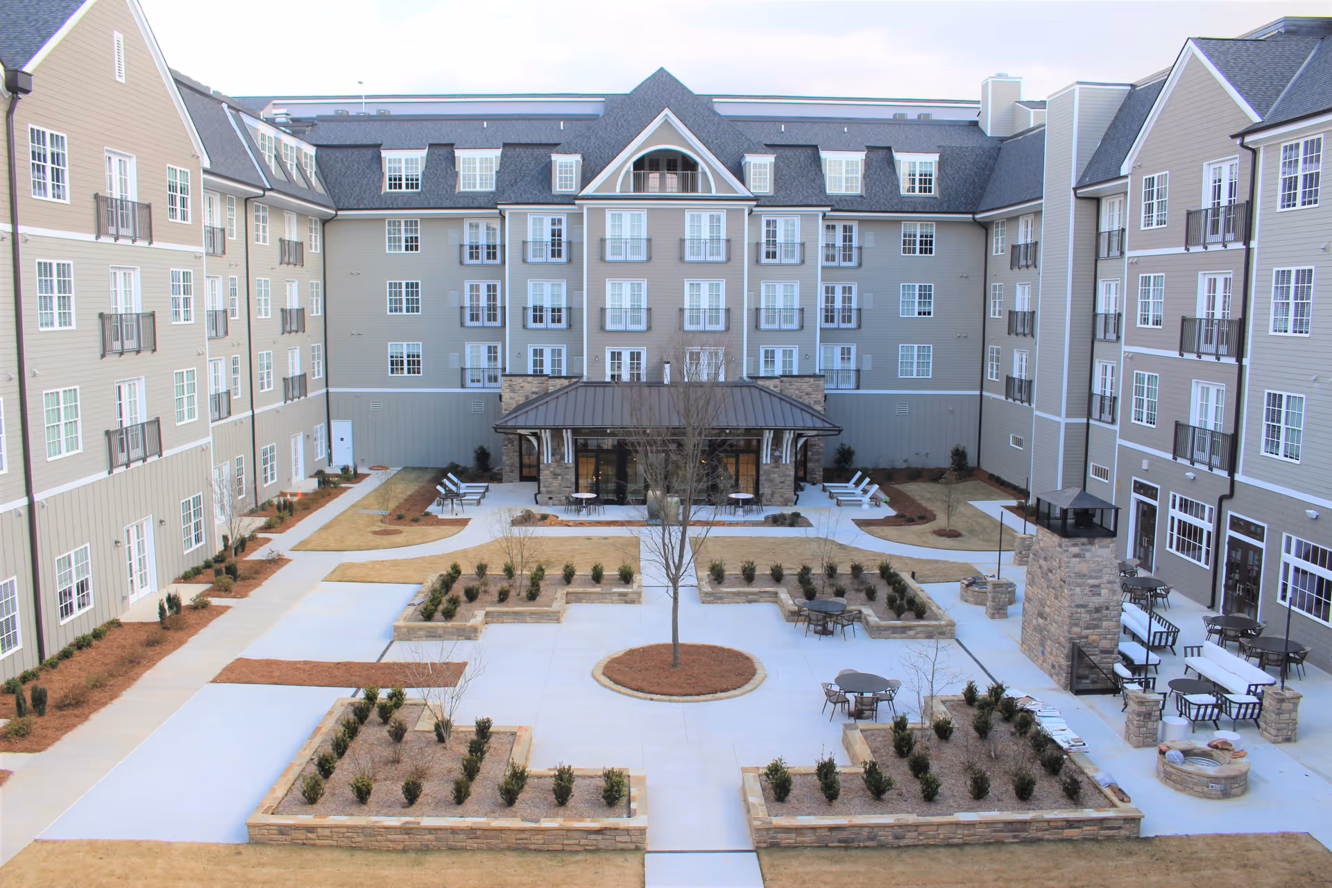 Outdoor courtyard area of a senior living facility with landscaped garden beds, paved walkways, seating areas with tables and chairs, lounge chairs, and a stone fireplace. The courtyard is surrounded by a multi-story building with numerous windows and balconies.